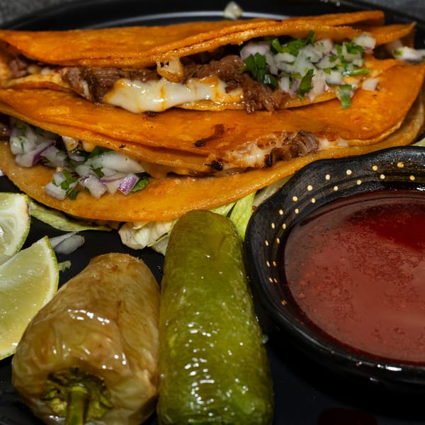 Two birria tacos with melted cheese, onions, cilantro, lime, and a side of consommé.