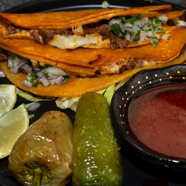 Two cheesy birria tacos with onions, cilantro, lime, peppers, and a side of consommé.