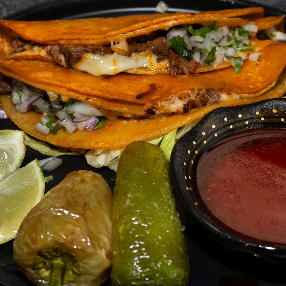 Two cheesy birria tacos with onions, cilantro, lime, peppers, and a side of consommé.