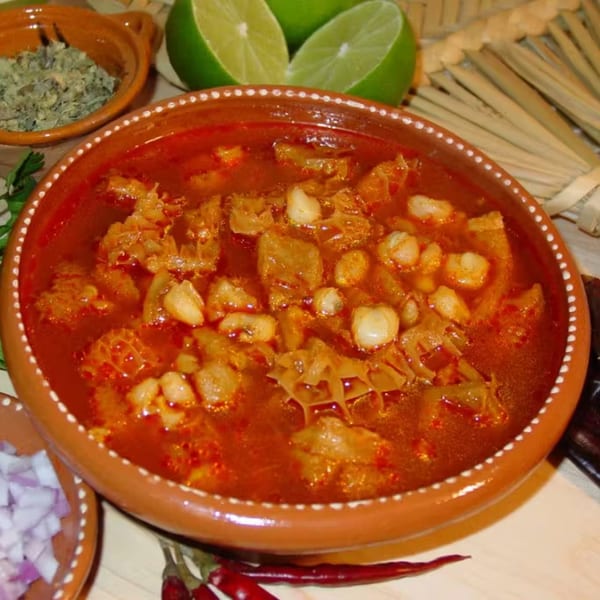 Menudo soup in a terracotta bowl with limes, dried herbs, and chopped onions on the side.