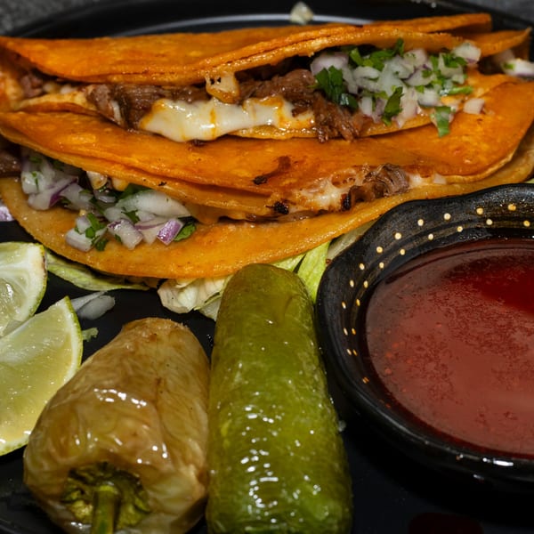 Two cheesy birria tacos with onions, cilantro, lime, peppers, and a side of consommé.