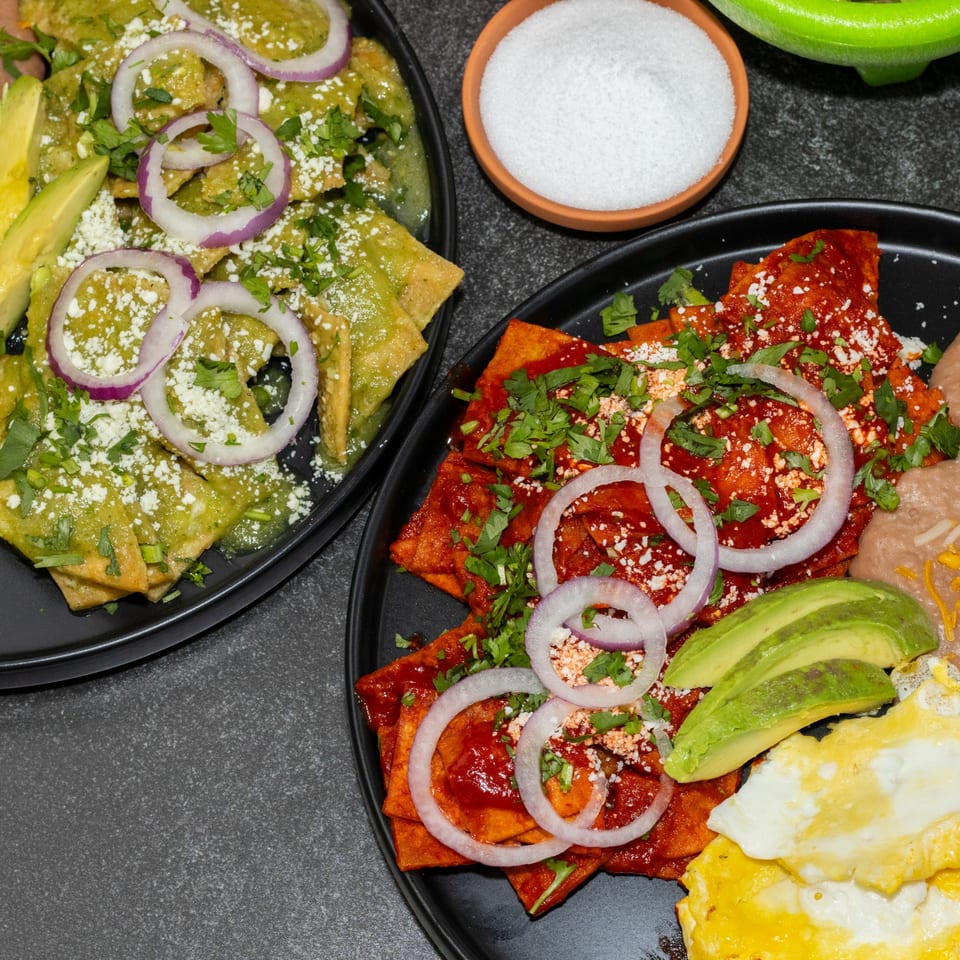 Two plates of chilaquiles, one green and one red, with avocado, onions, and cheese.
